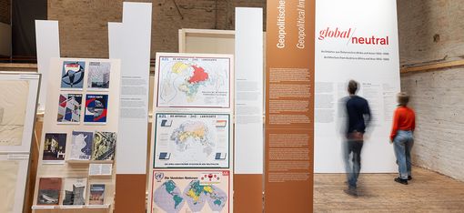 Very large pictorial elements stand on a wooden floor. In the background on the right, two people are reading a text.