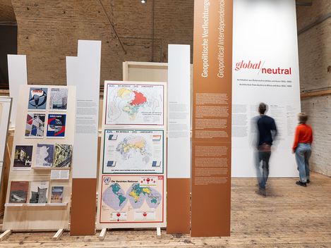 Very large pictorial elements stand on a wooden floor. In the background on the right, two people are reading a text.