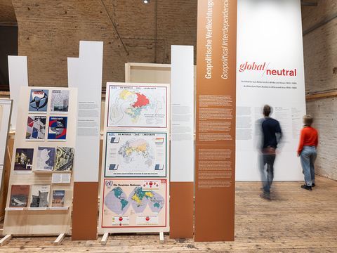 Very large pictorial elements stand on a wooden floor. In the background on the right, two people are reading a text.
