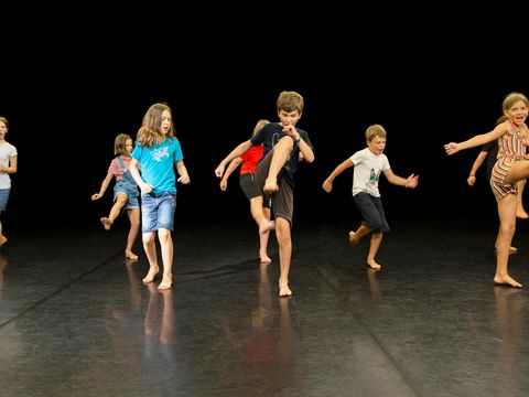 Several children are standing on a black dance floor, kicking their legs.