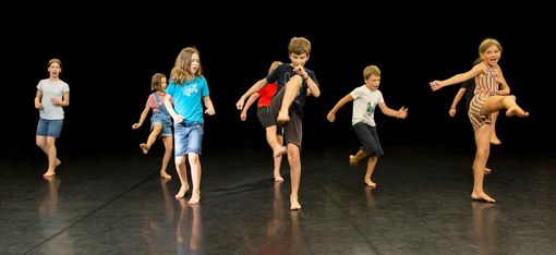 Several children are standing on a black dance floor, kicking their legs.