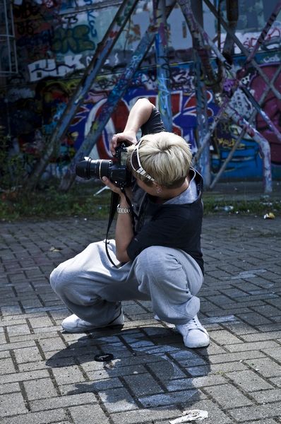 Person with short blonde hair crouches on paved ground holding a camera to take a photo.