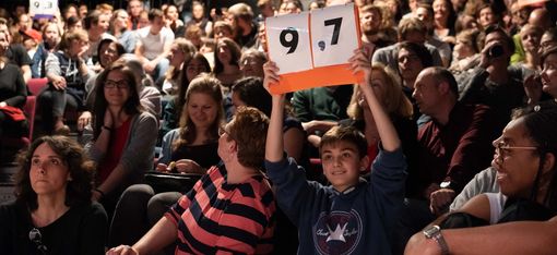 Publikum in einem Saal hält Wertungstafeln mit den Zahlen '9,7' hoch
