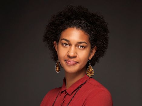 Frau trägt ein rotes Hemd mit langen Ärmeln und bunte Ohrringe und steht mit verschränkten Armen vor dunklem Hintergrund