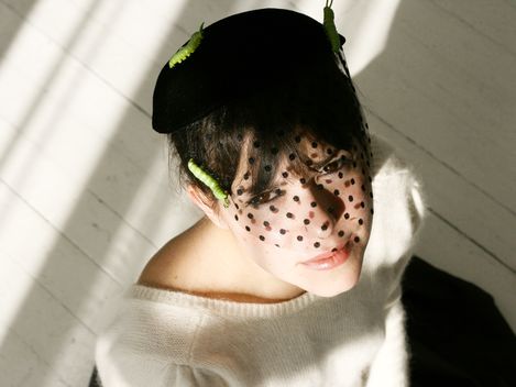 Woman wearing a black hat with a black net veil looking up while sitting on a white floor with shadow lines.