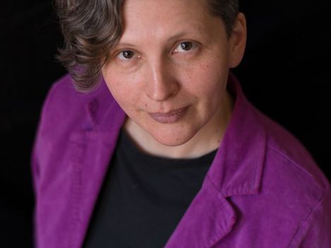 Woman with short wavy hair wearing a black top and a purple blazer against a black background.