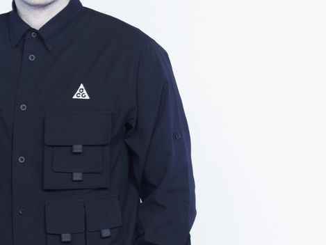 Man wearing a dark shirt with multiple pockets and a triangular logo on the chest against a light background.