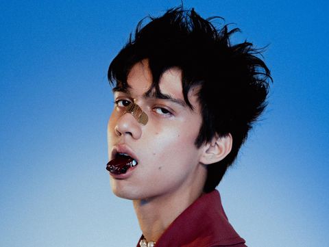 Person with tousled hair wearing a red blazer and pearl necklace against a blue background.
