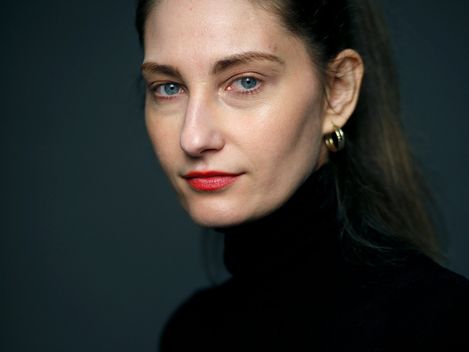 Woman with pulled-back hair, red lipstick, and black turtleneck against dark background