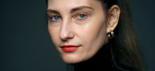 Woman with pulled-back hair, red lipstick, and black turtleneck against dark background