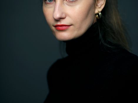 Woman with pulled-back hair, red lipstick, and black turtleneck against dark background