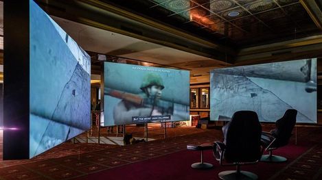 Three large screens showing historical black-and-white photos of soldiers in a room with chairs and carpeted floor.