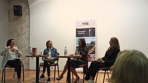 Four women sit on chairs around a table in a room with a poster on the wall behind them.
