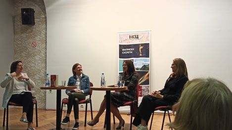 Four women sit on chairs around a table in a room with a poster on the wall behind them.