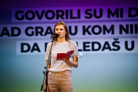 Woman standing at microphone holding a book in front of a wall with partially readable uppercase text.