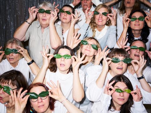 Fourteen people of various ages, dressed in white, are sitting close together in front of a glittering wall. They are holding green leaves up to their eyes.
