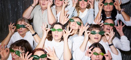 Fourteen people of various ages, dressed in white, are sitting close together in front of a glittering wall. They are holding green leaves up to their eyes.
