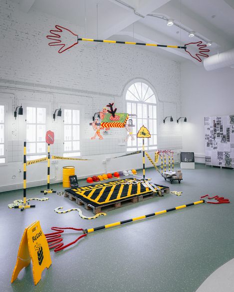 Art installation in a bright room featuring yellow-and-black warning stripes, large red hand shapes, and a figure hanging from a rope
