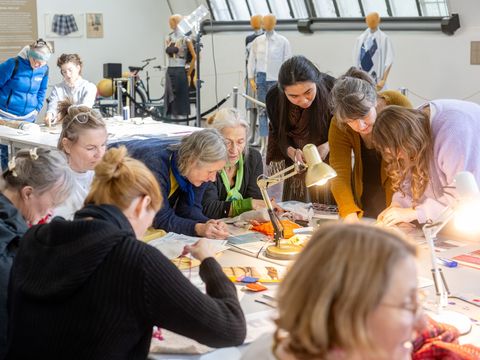Mehrere Personen sitzen an einem Tisch und arbeiten gemeinsam an Textilien in einem hellen Raum mit Schaufensterpuppen im Hintergrund.