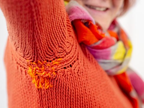 Frau mit roten Haaren, Brille und buntem Schal trägt einen orangefarbenen Pullover mit sichtbarer Reparatur am Ärmel