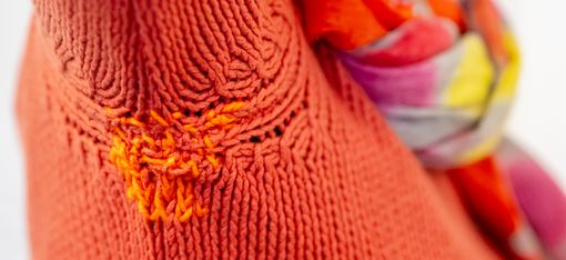 Frau mit roten Haaren, Brille und buntem Schal trägt einen orangefarbenen Pullover mit sichtbarer Reparatur am Ärmel