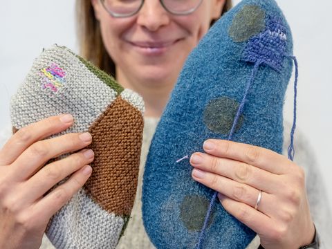 Frau mit Brille hält zwei handgefertigte, gestrickte oder gefilzte Objekte in verschiedenen Farben und Formen