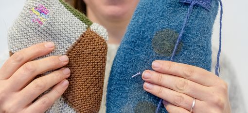 Frau mit Brille hält zwei handgefertigte, gestrickte oder gefilzte Objekte in verschiedenen Farben und Formen