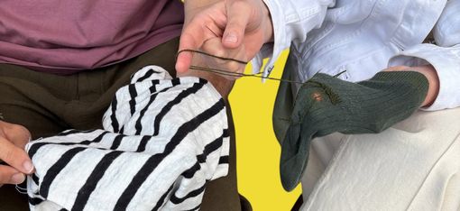 Zwei Personen sitzen nebeneinander, eine hält ein schwarz-weiß gestreiftes Kleidungsstück, die andere trägt eine helle Hose und braune Schuhe und hält eine grüne Socke mit einem Loch