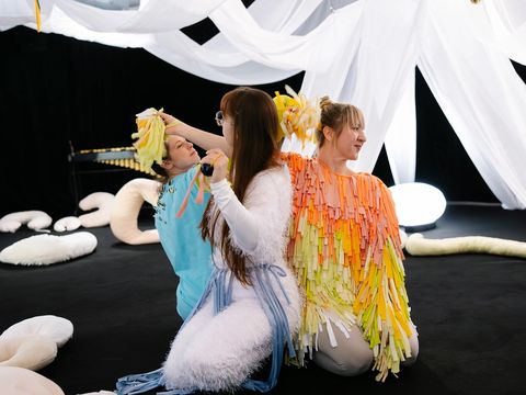 Three dancers in colourful, fluffy costumes are interacting with one another. They are sitting on a black floor, with white cloths hanging down from above and white cushions scattered around.
