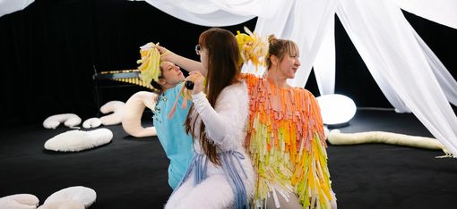 Three dancers in colourful, fluffy costumes are interacting with one another. They are sitting on a black floor, with white cloths hanging down from above and white cushions scattered around.