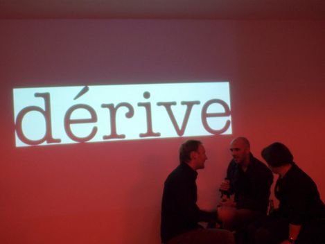 The word “dérive” is written on a large red canvas. Three people are standing in front of it.
