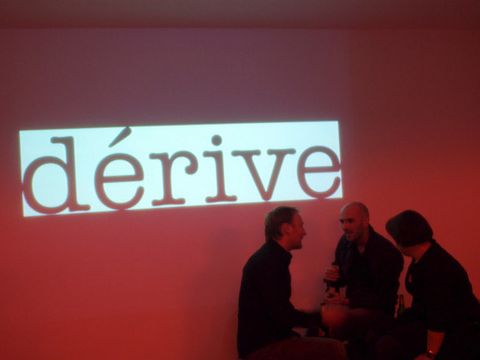 The word “dérive” is written on a large red canvas. Three people are standing in front of it.