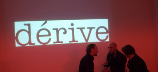 Der Schriftzug "dérive" ist auf einer großen, roten Leinwand zu sehen. Davor stehen 3 Personen.