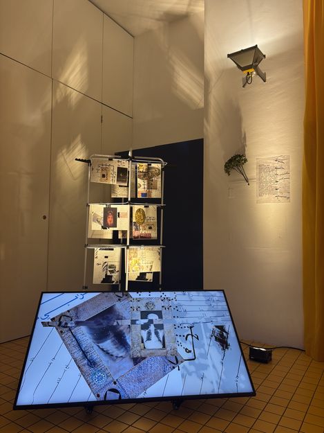 Kunstinstallation in einem Raum mit einer beleuchteten Tafel auf dem Boden, einem vertikalen Gestell mit angehängten Dokumenten und einer Wandlampe an der rechten Wand.
