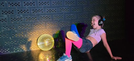 Ein Mädchen sitzt mit Kopfhörern auf dem Boden. Sie scheint sich zu unhörbarer Musik zu bewegen. Eine goldene Diskokugel, die neben ihr liegt, glitzert.