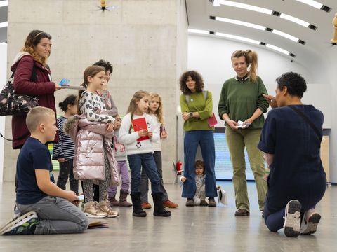 A person kneels in front of a group of children and adults in an exhibition room and explains something to them.