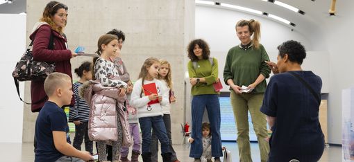 A person kneels in front of a group of children and adults in an exhibition room and explains something to them.