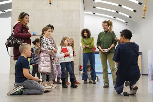 A person kneels in front of a group of children and adults in an exhibition room and explains something to them.