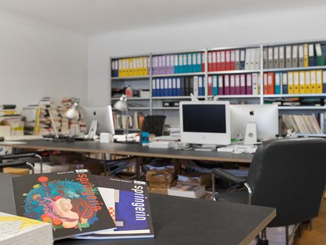 Büro mit mehreren Computern, Schreibtischen, Stühlen und einem Regal mit farbigen Ordnern im Hintergrund, im Vordergrund liegen zwei Zeitschriften auf einem Tisch