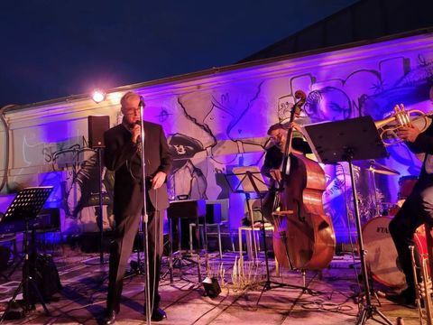 Four musicians perform on a stage in front of a wall with a large mural, including a singer with a microphone, a pianist, a double bassist, and a trumpeter.