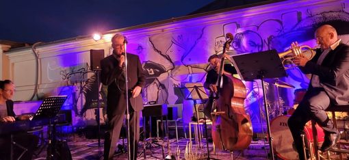 Four musicians perform on a stage in front of a wall with a large mural, including a singer with a microphone, a pianist, a double bassist, and a trumpeter.