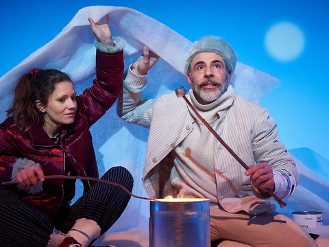 Two people are sitting in front of a makeshift campfire, holding marshmallows over it. They are holding a cloth over their heads.