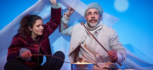 Two people are sitting in front of a makeshift campfire, holding marshmallows over it. They are holding a cloth over their heads.