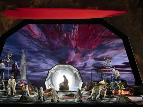 A theatrically staged set: In the center, a figure sits before a glowing, tunnel-like portal, surrounded by kneeling figures in bright robes. To the left and right, additional performers are grouped among symbolic objects and fire pits. In the background, a dramatic, vivid sky in shades of red and violet stretches out, lending the scene a mystical, ritualistic atmosphere.