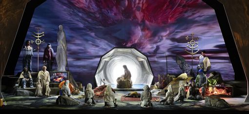A theatrically staged set: In the center, a figure sits before a glowing, tunnel-like portal, surrounded by kneeling figures in bright robes. To the left and right, additional performers are grouped among symbolic objects and fire pits. In the background, a dramatic, vivid sky in shades of red and violet stretches out, lending the scene a mystical, ritualistic atmosphere.