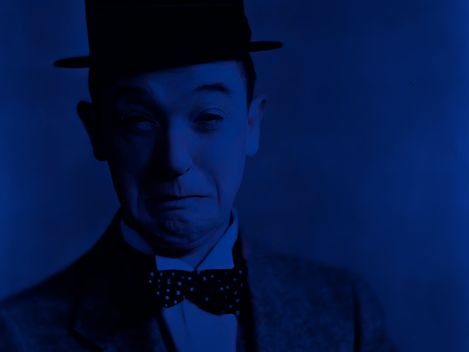 The image shows a portrait of Stan Laurel, captured in a highly stylized, monochromatic blue color scheme. He is wearing a dark suit with a bow tie and his signature round hat. His facial expression appears slightly uncertain or pensive—with furrowed eyebrows and a faint, hesitant smile.