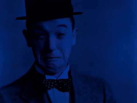 The image shows a portrait of Stan Laurel, captured in a highly stylized, monochromatic blue color scheme. He is wearing a dark suit with a bow tie and his signature round hat. His facial expression appears slightly uncertain or pensive—with furrowed eyebrows and a faint, hesitant smile.