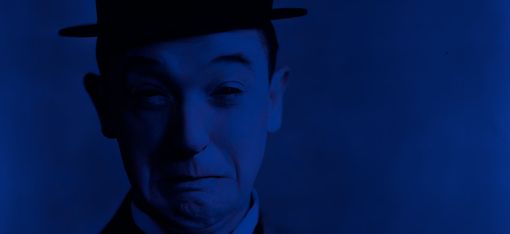 The image shows a portrait of Stan Laurel, captured in a highly stylized, monochromatic blue color scheme. He is wearing a dark suit with a bow tie and his signature round hat. His facial expression appears slightly uncertain or pensive—with furrowed eyebrows and a faint, hesitant smile.