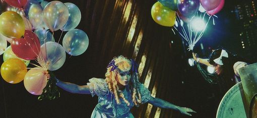 Two actresses wearing doll-like masks – one blonde, one brunette – both holding a bunch of balloons.