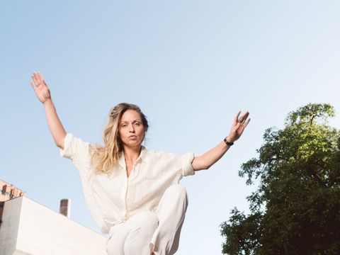 Person in heller Kleidung springt mit erhobenen Armen über ein Metallgeländer im Freien vor blauem Himmel